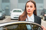 Woman in business suit and long hair parted in middle gets out of her car in parking lot. She holds the door and looks thoughtful, as if trying to remember the parking space she chose.