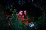 Two girls, lit in red, are seen sitting visibly scared under some lush vegetation.