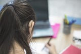 The back of a white female’s head with her brunette hair in a ponytail. We can see a skin tone behind the ear hearing aid on her right ear. She sits at a desk with a computer and paper in front of her, she is writing on the paper with a pen.