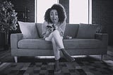 Black woman on a sofa with phone in one hand, the other hand is pressed against her face in an expression of horror and surprise.