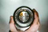 A hand holds a camera lens up against a blurred and indistinct background. In the middle of the lens the image is a sharp, well-defined picture of a city street.