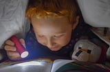 Young boy eager to learn using a flashlight and reading a book under his bed covers