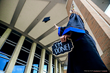 A student in a graduation gown tossing up their hat with a sign, “I’M DONE!”