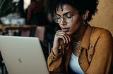 Woman with curly hair, wearing glasses sitting at a coffee shop looking at laptop.