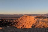 A picture of a desert landscape at sunrise. A large plateau is center of frame, with a craggy and dramatic canyon off to the left. The differences in elevation are so dramatic that the terrain looks almost like waves in a stormy ocean.