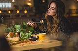 A woman with long brown hair, sitting at a wooden table at a restaurant. She is enjoying a bowl of pasta with something in a coffee mug. There is a viney plant next to her.