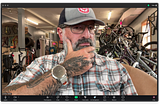 Screen capture of the Zoom user interface with the author looking at himself. He’s wearing a hat and glasses. His right hand is on his bearded chin, and his head is slightly turned, with his eyes looking directly into the camera. He’s in the thinking position. The tattoo on his hand is being primently displayed for all on Zoom to see.