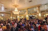 [ID: A room in the Austin Club packed with screenwriters of various genders and skin tones. The room has a white ceiling, cream colored arches, and a chandelier.]