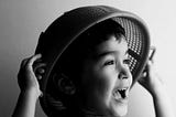 A greyscale photo of a boy holding a strainer over his head.