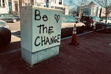 Photo looking towards the road, of a white electrical box on the  side of a red brick paved sidewalk, spray-painted with black lettering “Be the change” and a heart. Two orange traffic cones are nearby, in front of parked cars.