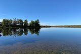 A picture of a calm and clear lake in Maine with the reflection of trees in the water.