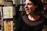 Caitlin Moran in three quarter profile with open grin, holding a glass of water in front of books, wearing black smock top