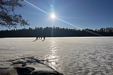 3 persons ice-skating on a frozen lake. Blue skies, the sun is shining. Frozen white lake with glimmering sun over the ice. Forests and trees in the surroundings.