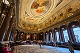 A large hall with marble pillar all around and a ceiling with an oval central painting, and rectangular ends with paintings in them. Arches embellished with gold behind an upper balcony are above the pillars.