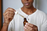 A person wearing a textured, off-white shirt is holding a small amber glass dropper bottle in one hand and a dropper filled with a light-colored liquid in the other. The dropper is being removed from or lowered into the bottle, and a droplet is visible at the tip. The person’s face is partially visible, softly focused in the background, while the hands and bottle are in clear focus in the foreground.