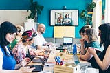 7 members of the Clearleft team sit around a large table in their studio having a meeting. A further 4 team members have joined remotely and can be seen on the TV in the background.