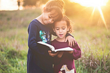 Two sisters standing in a sunny field, reading a book
