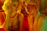 Two young blond women, Violet and Eddie, are sitting at a table in a red-lit bar. A magazine lays open on the table. Violet is staring fondly at Eddie who peers out at the viewer in thought.