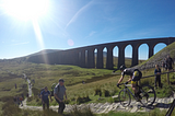 A cyclist is descending a man-made stone path in the Yorkshire Dales. There is a long stone viaduct in the background. The sky is blue and there are several spectators randomly spaced out along the route which goes down a hill lush with green moss.