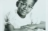 Little black girl wearing glasses, seated at desk, smiling