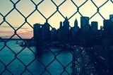 The view of downtown city that I snapped while walking over the Manhattan Bridge during sunset. The sky is yellow, the water beside the highway dark blue, the city buildings darkening with the sky. There are many cars on the highway with headlights facing the direction of the camera, and others facing in the opposite direction.