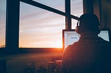 man sitting facing back in front of laptop next to window as the sun sets