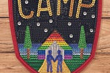 The cover of Camp by LC Rosen. The background is lightly colored wood planks, and a large sequined badge takes the foreground. Camp is spelled out in gold sequins in a starry night. On the gorund level, two blue-sequined figures hold hands in front of a gay rainbow colored building.