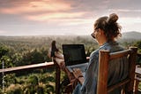 Woman sitting on chair on deck overlooking wilderness. She has feet on rail while working on laptop.