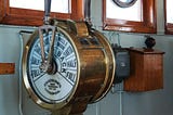 A brass ship’s engine order telegraph (ship’s telegraph) mounted on a pedestal in what appears to be a ship’s bridge or wheelhouse. The circular brass instrument has a white face with black markings showing different speed commands including “FULL,” “HALF,” “SLOW,” and “BACK.