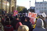 Photograph of students protesting in Westminster, 10 November 2010. Crowd with placards, seen from the back. In the distance the Houses of Parliament, sun reflecting in a window.