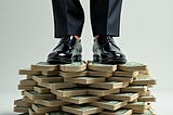 A pair of men’s legs in expensive shoes and suit trousers, standing on top of a large stack of paper money.