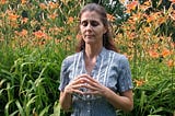 A woman kneeling with her fingertips touching. Her thumbs are against her chest and her elbows are relaxed against her body. She’s wearing a black and white vertically striped shirt in front of a patch of orange flowers.