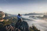A photographer wearing a hat and blue waterproof looking through their camera while sitting on top of a mountain surrounded by clouds