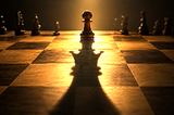 A worn wooden pawn stands on the far edge of a chessboard, backlit by golden light, casting a long shadow toward the viewer in the unmistakable shape of a queen.