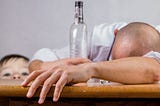 Toddler peeking up from under a table seeing a man holding an empty bottle of alcohol and shot glass with his head down.