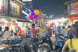 Motorcycles are parked in the center of this picture. Someone’s selling socks off one of the motorcycles. A toy seller sells balloons and toys. Clothes shops display their wares, There are plenty of people in this picture. Some people are walking around with their motorcycle helmets on.