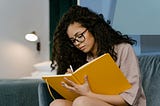 Woman with glasses and curly brown hair sitting on a couch writing in a yellow journal or notebook