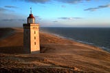 A light house standing in a sand dune is illuminated by a setting sun.