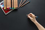 An image of a box of colored pencils on a black background. A hand holds one of the pencils near the middle.