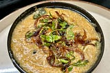 On a white plate, a black bowl holds a portion of brown-colored stew with shredded meat, garnished with caramelized onions, coriander leaves, and mint leaves.