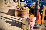 image shows a scraggly haired toddler in a pink coat on a brick sidewalk, playing with a metal keg that is next to a black lamppost. In the background, there is a brick planter with colorful flowers