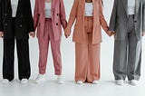 Four women standing side by side, holding hands. They are dressed in stylish, oversised suits in various colours: black, pink, peach, and grey. The image is cropped to show their outfits from the shoulders down, highlighting their coordinated, fashionable attire and unity.