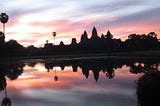 A silhouette of Angkor Wat, Cambodia, at sunrise. The structure and sky are perfectly reflected in a lake. The sky is streaked with fine clouds in pale blue, white, peach, yellow and purple.