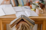 A person is sitting at a desk with books scattered on top. Their head is tipped back with an open book covering their face.