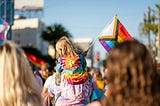 A young child wearing a tie-dye shirt and holding the Pride flag sits atop their parent’s shoulders at a Pride parade