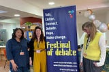 The three researchers standing next to the Festival of Debate poster, which is blue with yellow writing. Yinka is wearing blue and Christina & Louise are wearing yellow.