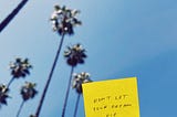 A yellow sticky note held up against blue sky with palm trees that reads “don’t let your dream die.”