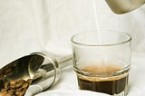 Coffee beans scoop placed on white sheet with a small quantity of beans inside and glass mug having espresso is also kept here ,someone pouring milk from milk frothing stainless steel jar into the mug ,background is white to enhance the colors.