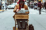 Image of busy city street, and in the middle of the street is a man at a desk with a sign reading “Poet for Hire”