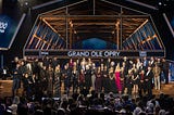 A large group of Grand Ole Opry members from multiple generations, including Bill Anderson, Connie Smith, Randy Travis, Reba McEntire, Carrie Underwood, and Carly Pearce, stand together on stage at the Opry House during the 100th Anniversary Celebration in 2025, beneath the Grand Ole Opry sign and wooden stage beams, honoring a century of country music tradition.